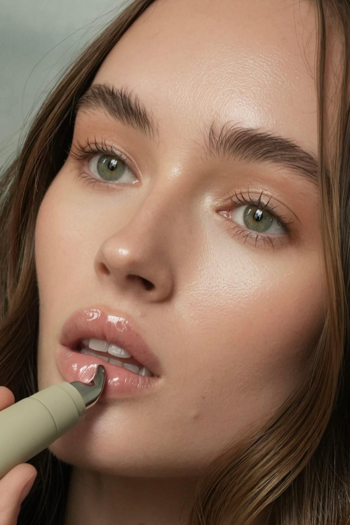 Close-up of a model applying league with a metal applicator, showing glossy pink lips against soft neutral lighting.